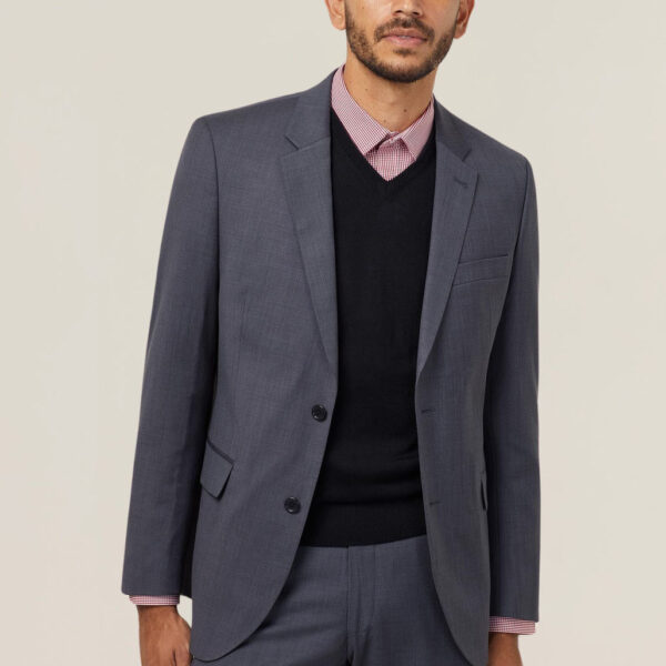 A person with short dark hair and a beard is wearing a dark gray suit, a black sweater, and a pink checkered shirt. They are standing against a plain light background, looking towards the camera with a neutral expression.