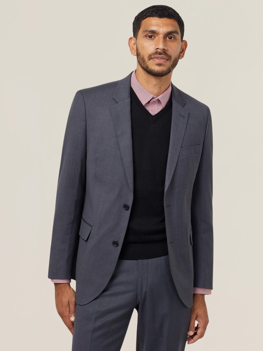 A person with short dark hair and a beard is wearing a dark gray suit, a black sweater, and a pink checkered shirt. They are standing against a plain light background, looking towards the camera with a neutral expression.
