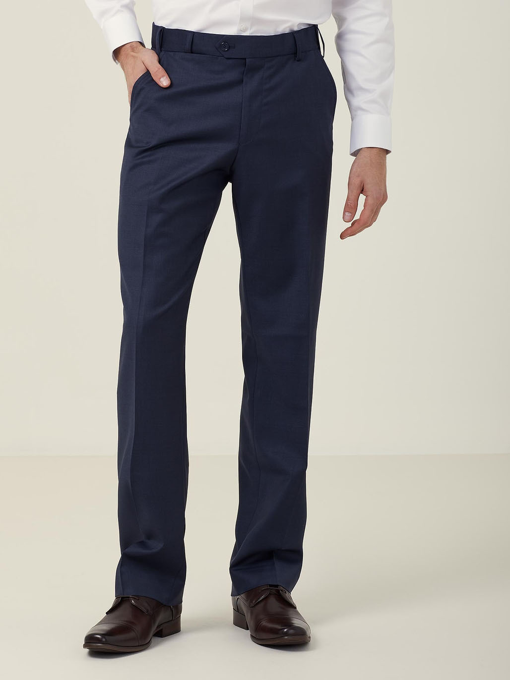 A person wearing a white dress shirt and dark blue formal trousers, standing with one hand in a pocket. The outfit is completed with dark brown dress shoes. The background is plain and neutral.