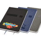 The Emz A5 Notebook collection features three hardcover options with fabric covers in black, blue, and gray. The black version has a fruit logo and The Fruit Basket text. All notebooks include a ribbon bookmark and an elastic band closure for convenience.