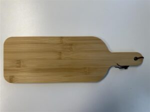 A rectangular wooden cutting board with a handle rests on a white surface, ideal as a promotional item. The handle features a hole with a black string looped through it. Natural wood grain adds texture to the board, reminiscent of outdoor experiences in rustic settings.