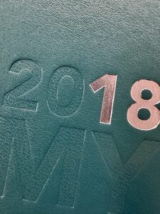 Close-up of a textured teal surface showcasing embossed silver numbers 2018, achieved through foil debossing. Parts of the other embossed letters MY elegantly integrate into the teal surface, adding depth to this intricate design.