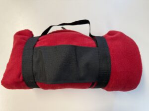 A rolled-up red fleece blanket, ideal for outdoor experiences, secured with black straps and a handle, sits invitingly on a white surface. Perfect as promotional items to enhance your adventures.