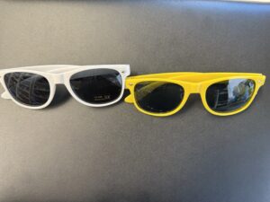 Two pairs of sunglasses are displayed on a black surface: one white with dark lenses, the other yellow. Both showcase a classic wayfarer-style frame, perfect promotional items for enhancing outdoor experiences with eye-catching flair.