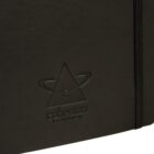 Close-up of a black notebook with an embossed logo on the front cover. The logo features a stylized planet with rings and the text cohesion branding beneath it. A black elastic band is visible on the right edge of the notebook.