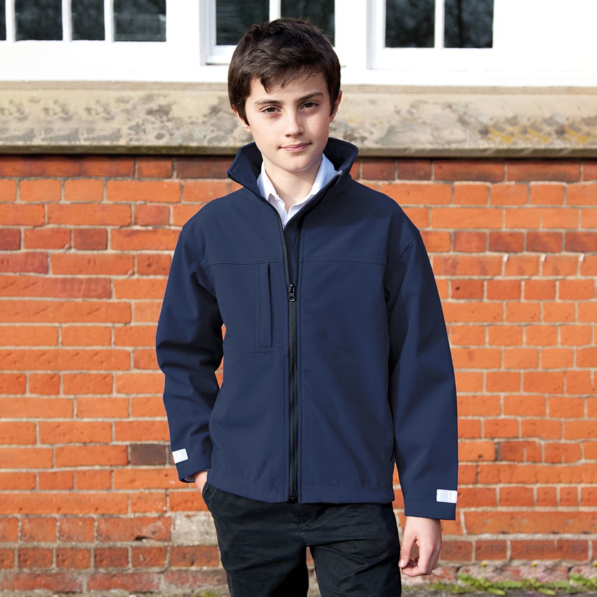 A boy in a navy jacket stands in front of a red brick wall and white windows.