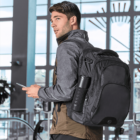 Man with a black backpack holding a phone, standing indoors by large windows.