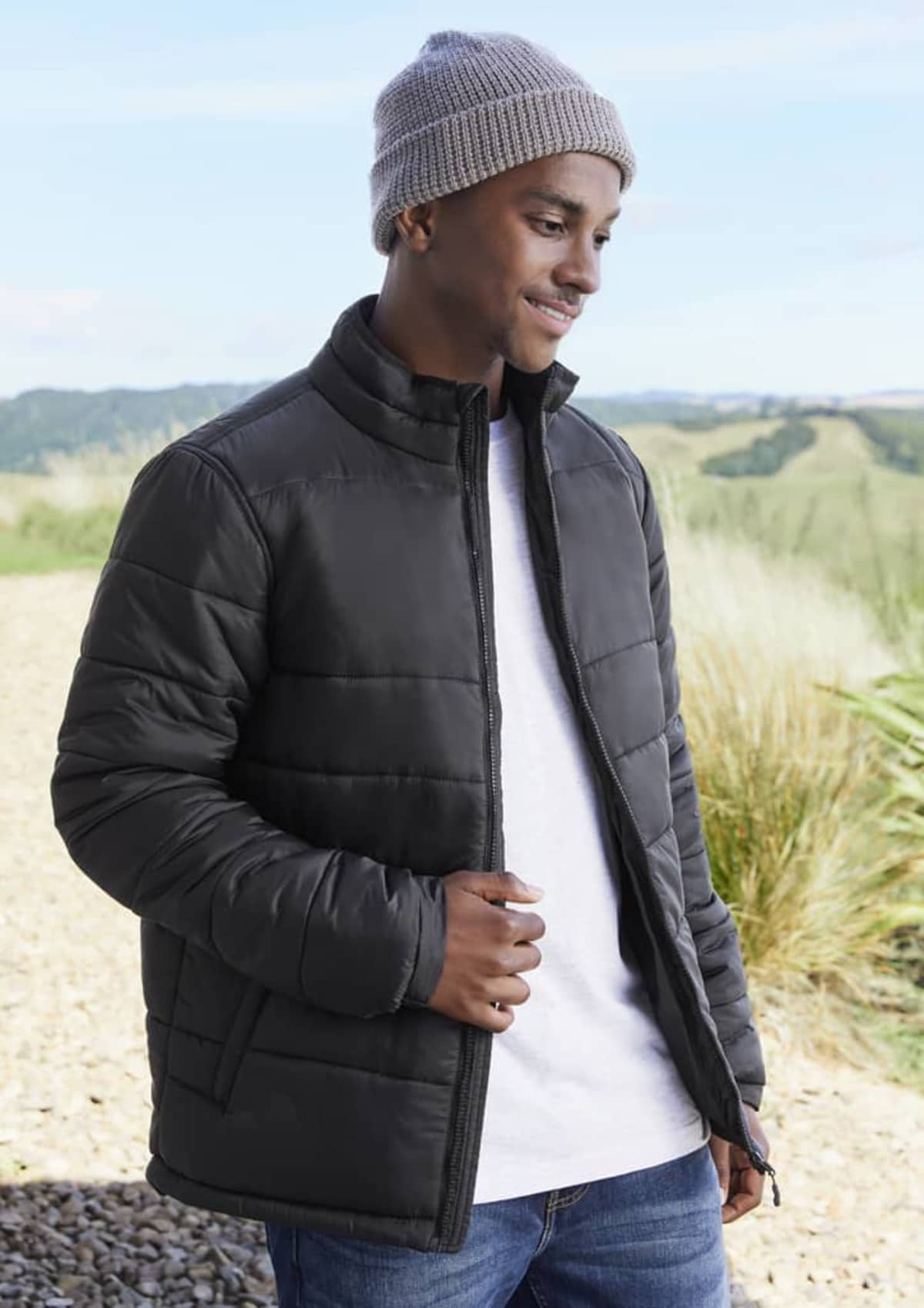 Man in a gray beanie and black puffer jacket standing outdoors, smiling.