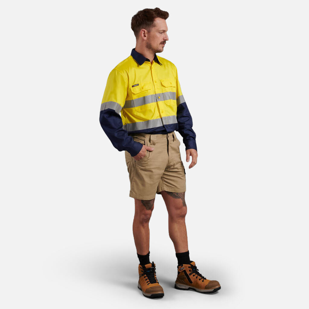 Man in Explorer Shield shirt, khaki shorts, and boots, hand in pocket.