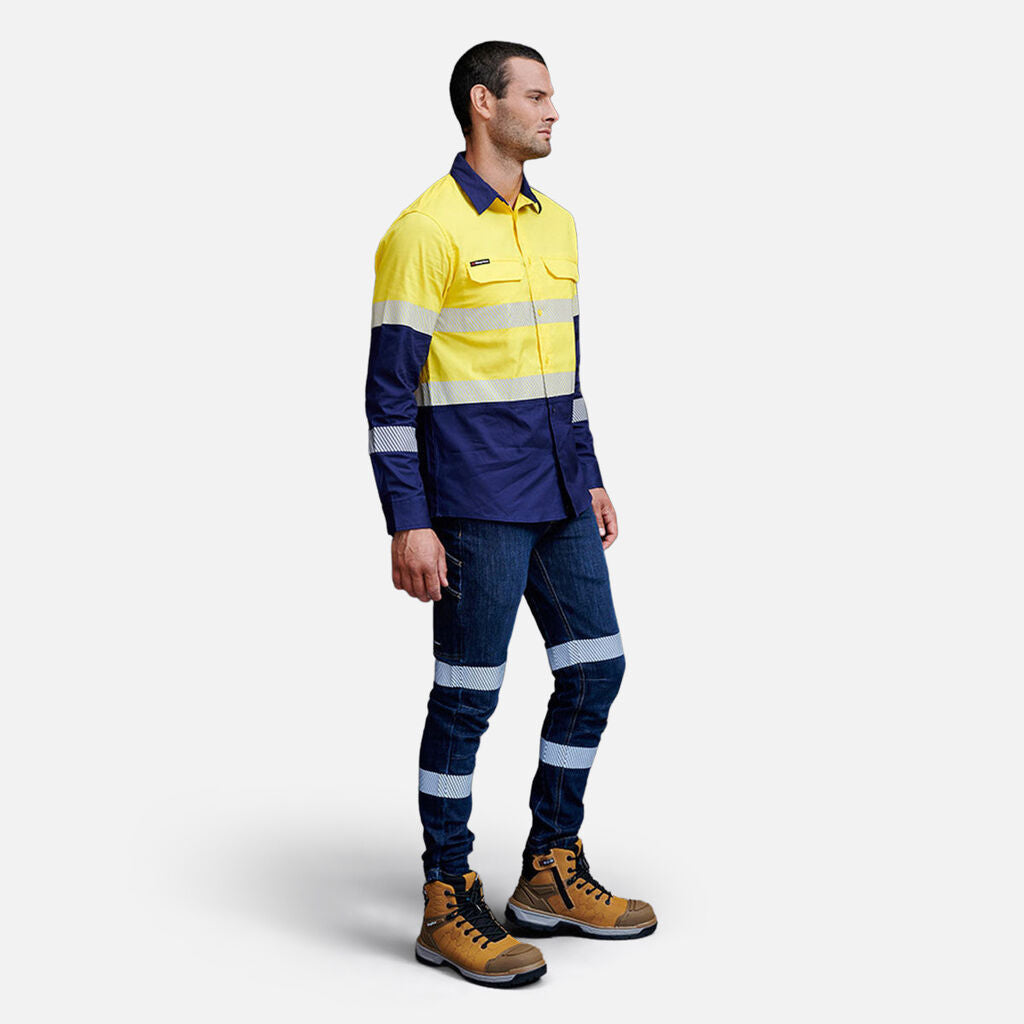 Against a gray backdrop, an individual wears a yellow and navy high-visibility work shirt with reflective bands, navy pants with reflective stripes, and brown work boots. In their hand is an Emz A5 Notebook, poised to jot down important details.