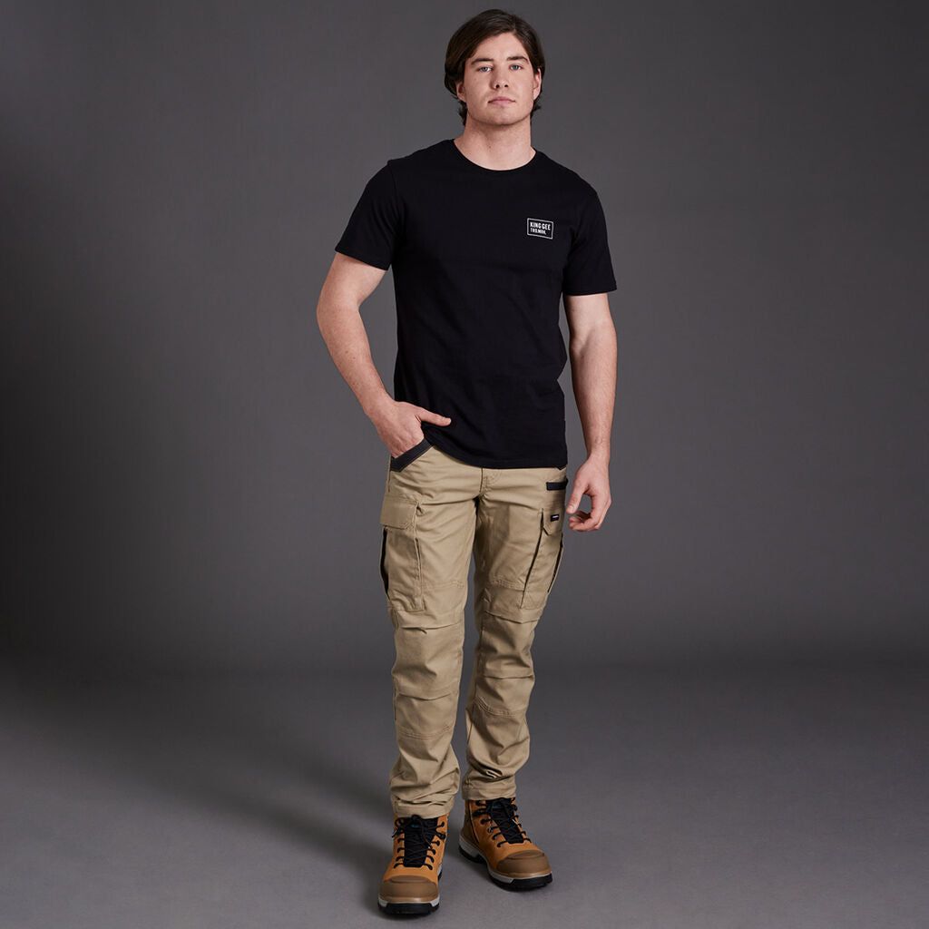 Man in Explorer Shield black tee, khaki cargo pants & work boots stands against gray background.