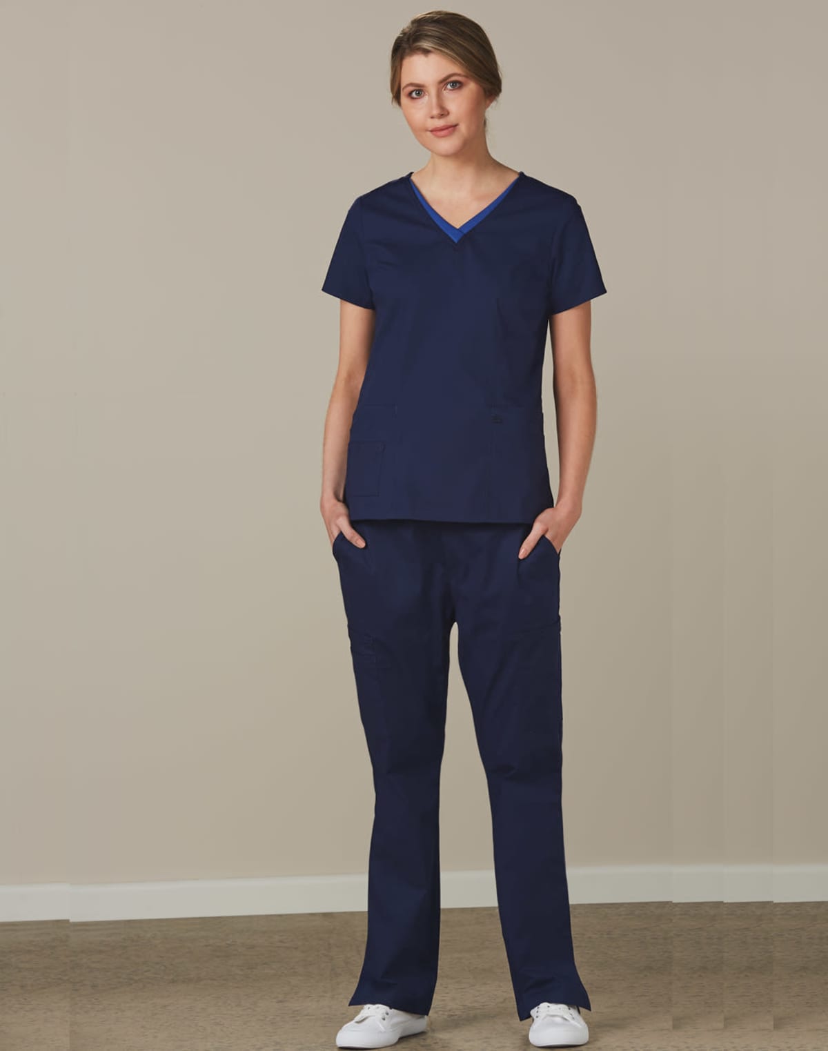 Person standing in navy blue scrubs and white shoes against a plain beige background.