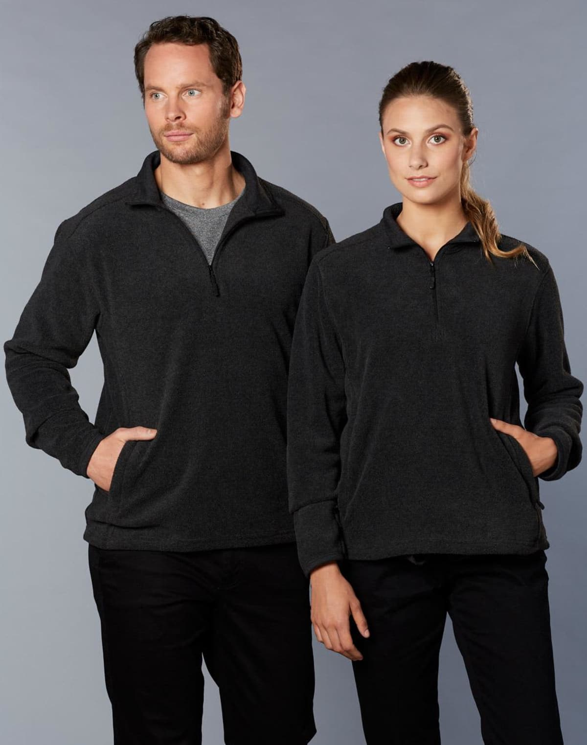 Man and woman in matching dark quarter-zip fleece tops with hands in pockets, against gray background.