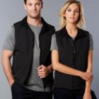 Two people in grey shirts and black vests standing side by side against a grey background.