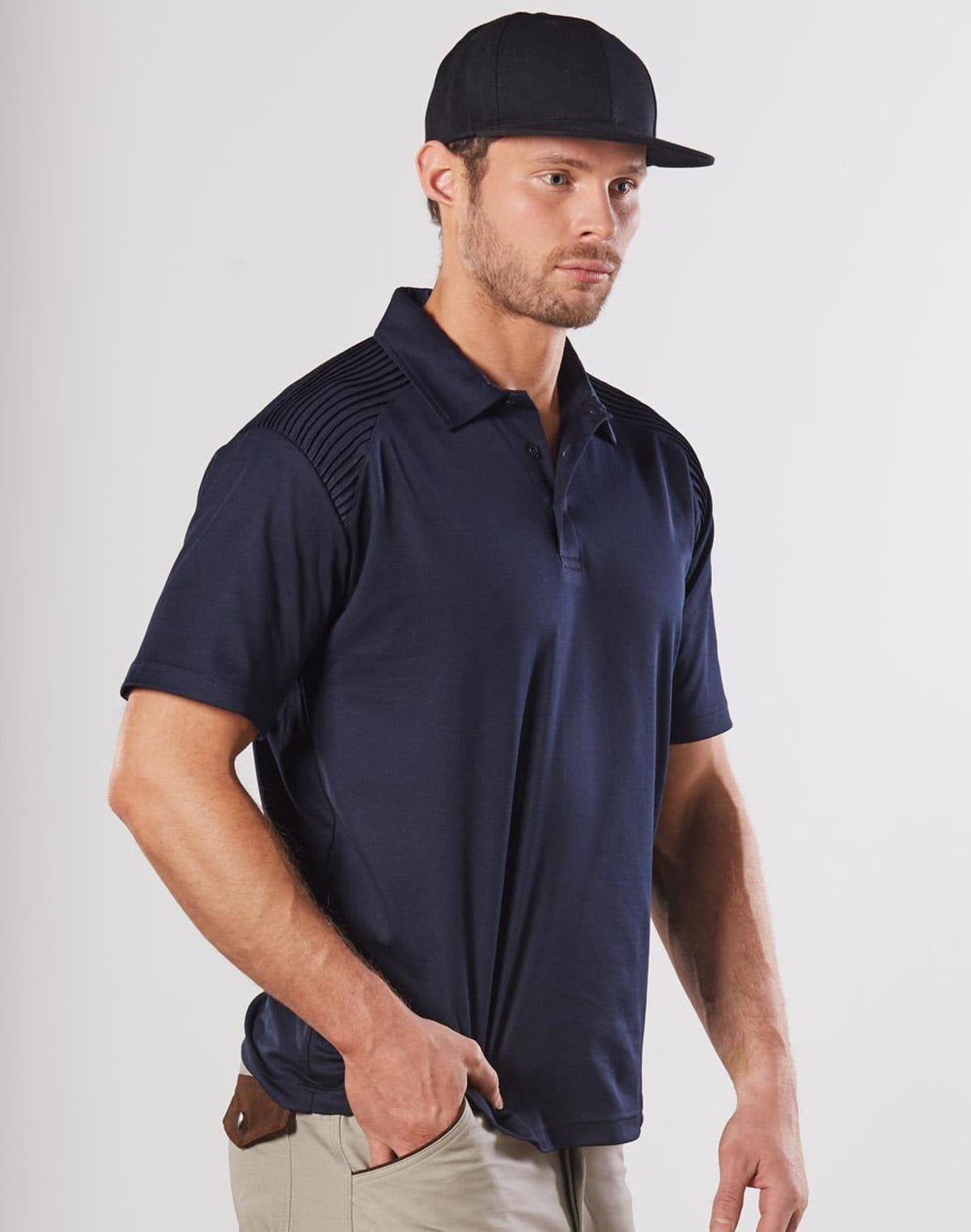 Man in a navy polo shirt, beige pants, and a black cap standing against a plain background.