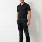 Man in black workwear and tan boots standing against a white brick wall.