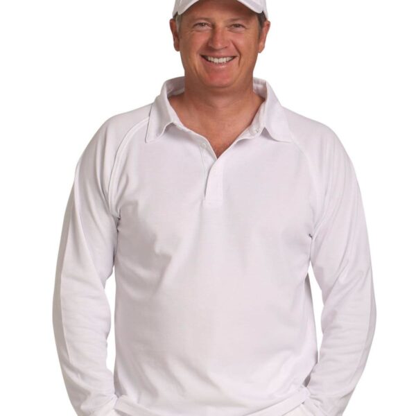 Man in a white cricket uniform and cap smiling with hands in his pockets.
