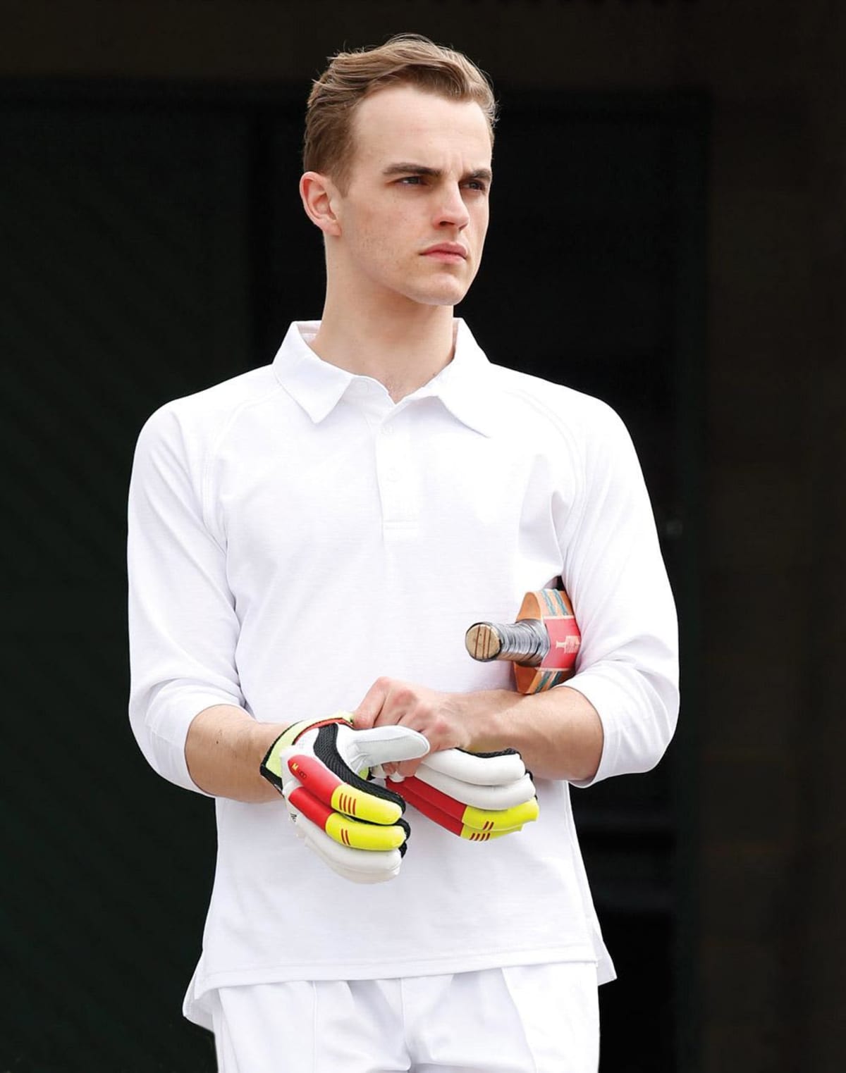 Man in white cricket attire holds gloves and bat, looking to the side.