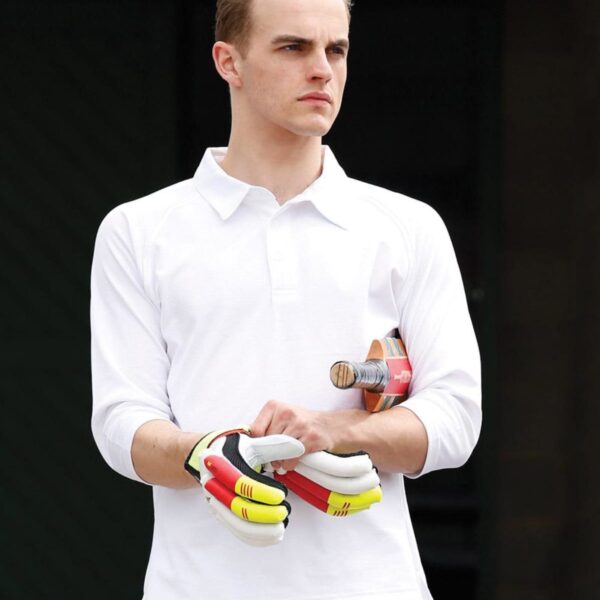 A cricket player in white clothes holds gloves and a bat under his arm.
