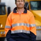 Man in a high-visibility orange work jacket standing in front of yellow trucks.