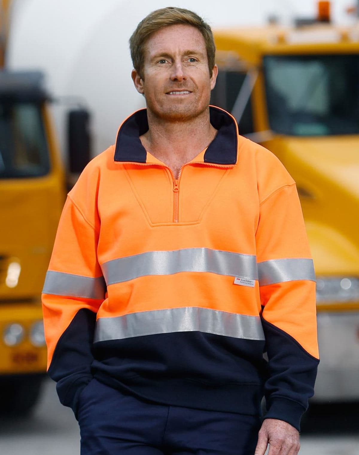 Man in a high-visibility orange work jacket standing in front of yellow trucks.