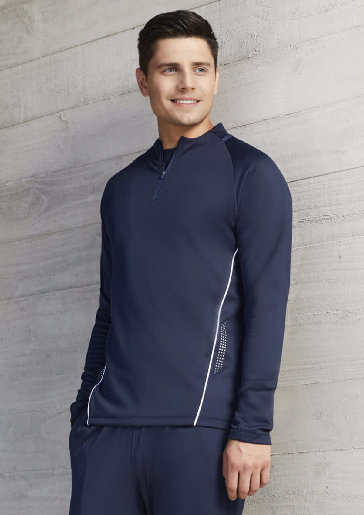 Man in navy blue athletic jacket standing against a light gray concrete wall, smiling.