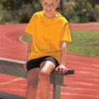 Smiling child in a yellow shirt sits on a bench by an outdoor running track.