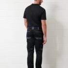 Man in black polo and cargo pants, facing away from camera, standing by a white brick wall.
