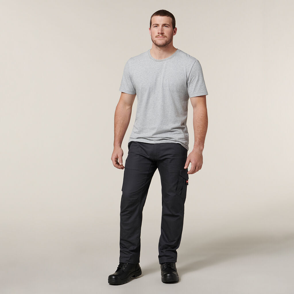 A man stands against a plain background, dressed in a light gray T-shirt, dark gray cargo pants, and black boots. In his hands, he gently holds an Emz A5 Notebook by his side while gazing slightly away.