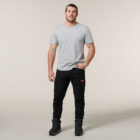 The person stands against a neutral background, wearing a light gray Emz t-shirt and black pants with pockets. They have short hair, black shoes, one hand casually in their pocket while holding an Emz A5 Notebook in the other, looking directly at the camera.