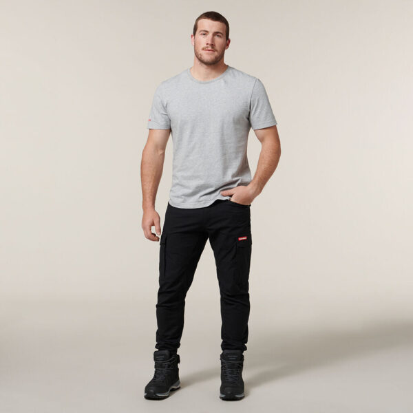 The person stands against a neutral background, wearing a light gray Emz t-shirt and black pants with pockets. They have short hair, black shoes, one hand casually in their pocket while holding an Emz A5 Notebook in the other, looking directly at the camera.