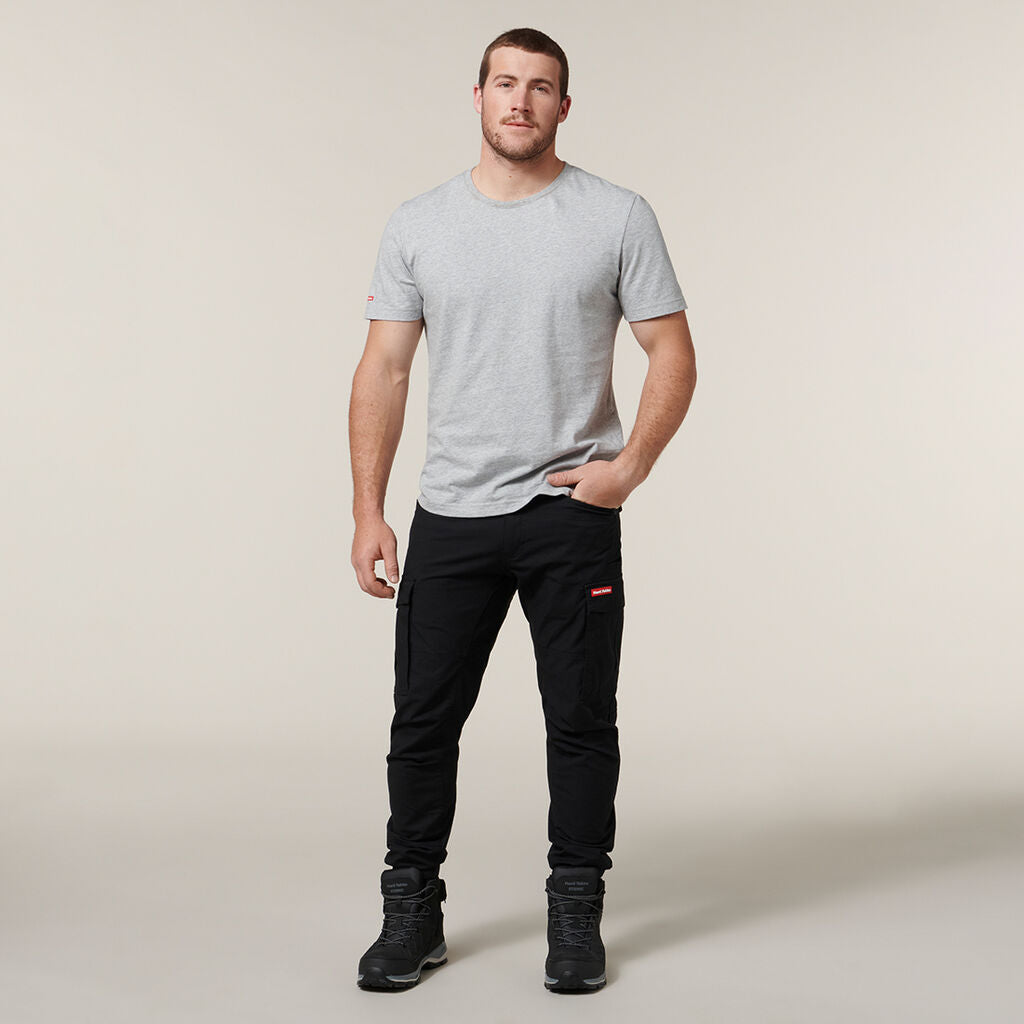 The person stands against a neutral background, wearing a light gray Emz t-shirt and black pants with pockets. They have short hair, black shoes, one hand casually in their pocket while holding an Emz A5 Notebook in the other, looking directly at the camera.