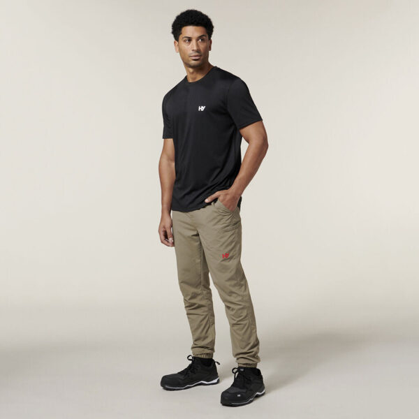 Wearing a black t-shirt and khaki pants, a person poses against a neutral background, holding an Emz A5 Notebook in one hand. They have short hair and black shoes.