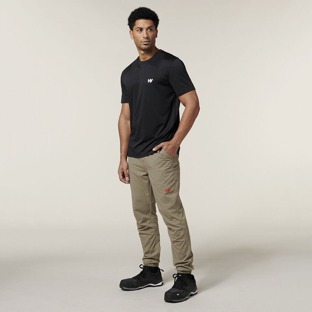 Wearing a black t-shirt and khaki pants, a person poses against a neutral background, holding an Emz A5 Notebook in one hand. They have short hair and black shoes.