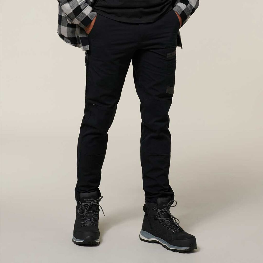 A person wearing black cargo pants, a black shirt, and a plaid jacket stands with hands in pockets. An Emz A5 Notebook subtly peeks out. Theyre wearing black hiking boots with grey soles against a plain, neutral background.