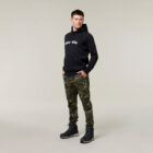 A man stands against a neutral backdrop, wearing a black hoodie with white text, camouflage pants, and black boots. An Emz A5 Notebook peeks from his pocket as he gazes off to the side, deep in thought.