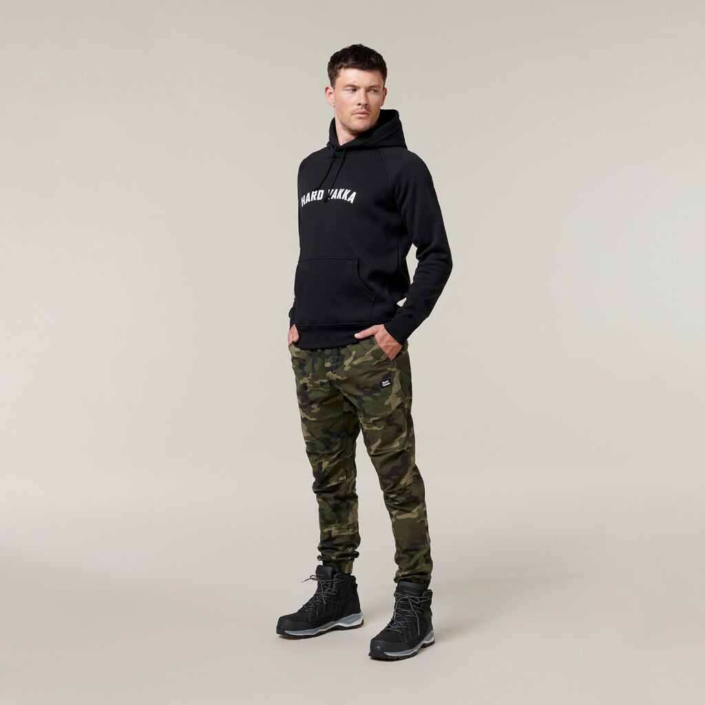 A man stands against a neutral backdrop, wearing a black hoodie with white text, camouflage pants, and black boots. An Emz A5 Notebook peeks from his pocket as he gazes off to the side, deep in thought.
