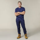 Emz stands with arms crossed, dressed in a navy blue T-shirt and matching pants with light brown work boots. An Emz A5 Notebook peeks out of the pocket against the off-white background.
