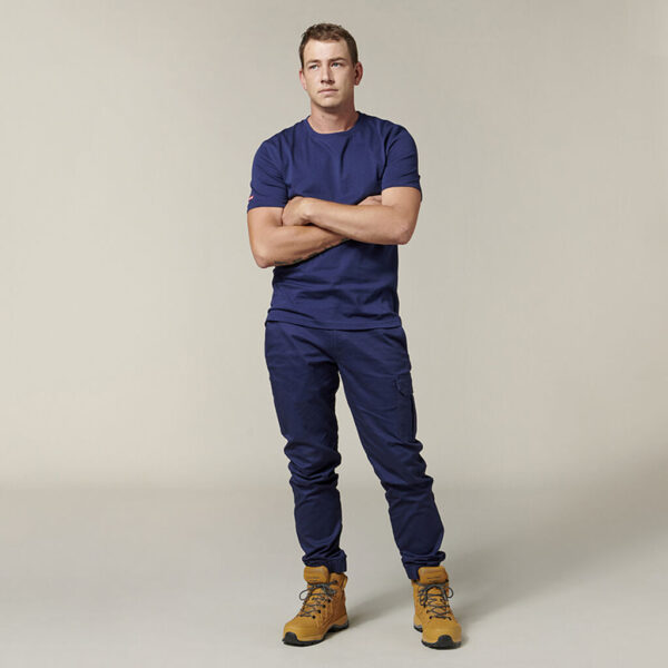 Emz stands with arms crossed, dressed in a navy blue T-shirt and matching pants with light brown work boots. An Emz A5 Notebook peeks out of the pocket against the off-white background.