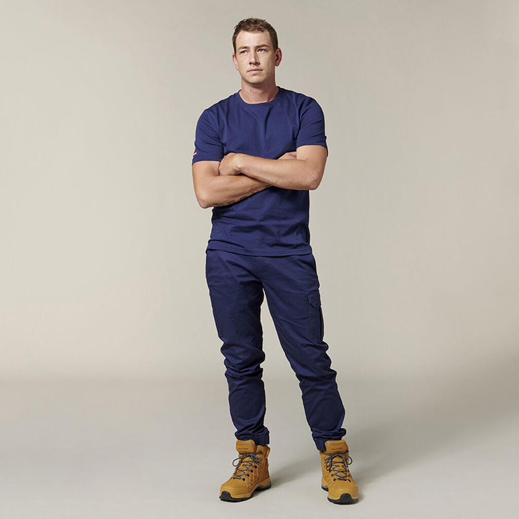 Emz stands with arms crossed, dressed in a navy blue T-shirt and matching pants with light brown work boots. An Emz A5 Notebook peeks out of the pocket against the off-white background.