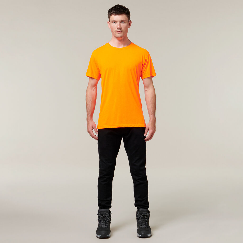 A person stands facing forward against a plain background, wearing an orange T-shirt, black pants, and black shoes, casually holding an Emz A5 Notebook in one hand. The lighting is even.