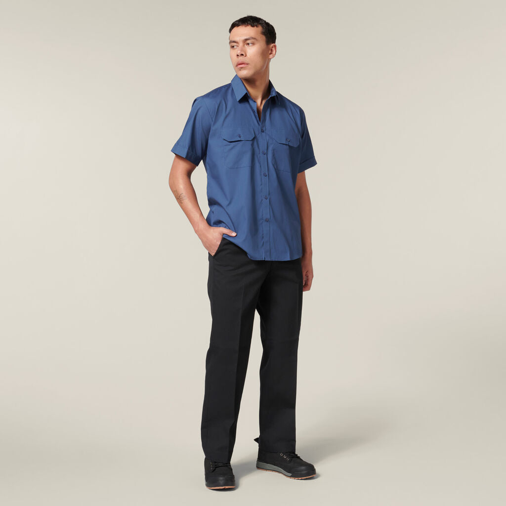 A person stands against a neutral background, wearing a blue short-sleeve button-up shirt and black pants. Their left hand is in the pants pocket while they glance to the side. They hold an Emz A5 Notebook in their right hand, complementing their outfit with black shoes for a smart-casual look.