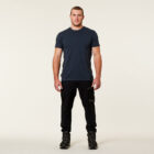 A man stands against a plain background, wearing a dark t-shirt, black cargo pants, and firmly planted boots. He faces the camera with a neutral expression and hands by his sides.