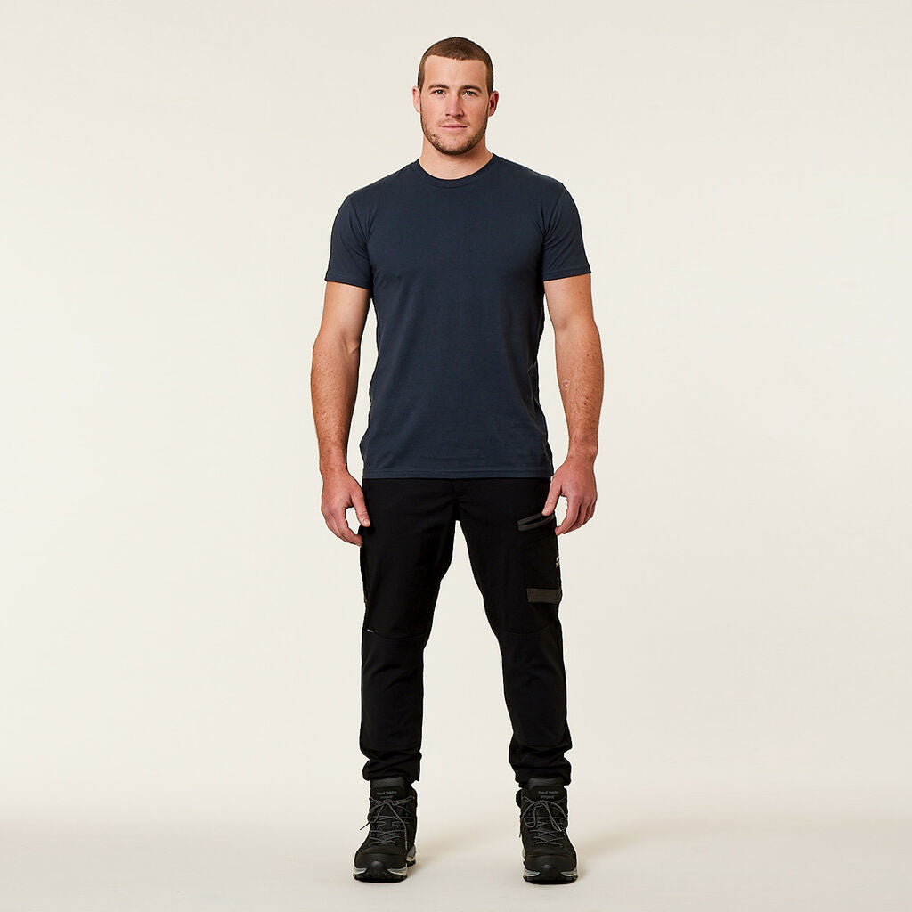 A man stands against a plain background, wearing a dark t-shirt, black cargo pants, and firmly planted boots. He faces the camera with a neutral expression and hands by his sides.