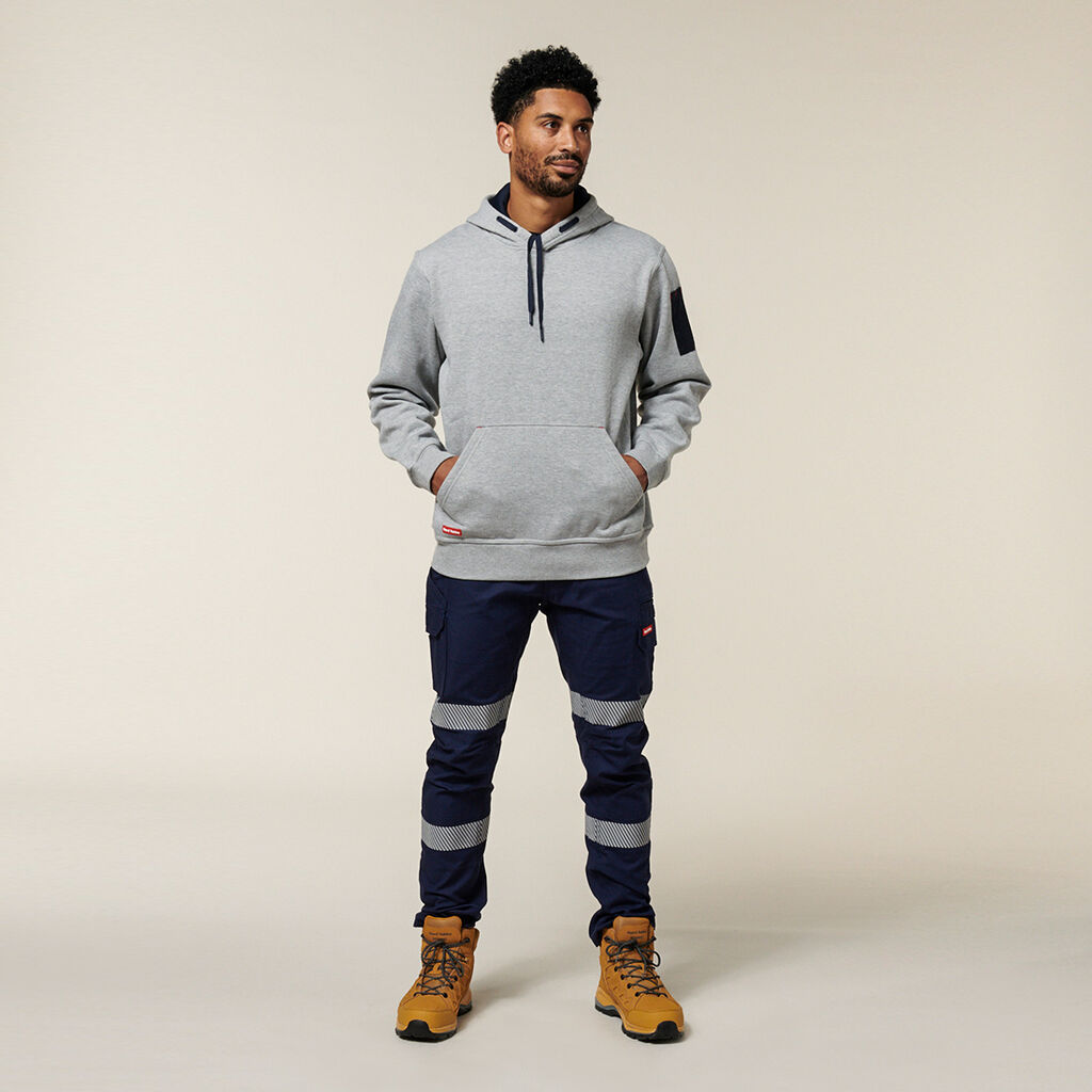 A person in a gray hoodie, navy cargo pants with striped accents, and tan boots stands against a neutral background, hands casually in pockets as they glance aside, seemingly contemplating notes for their Emz A5 Notebook.