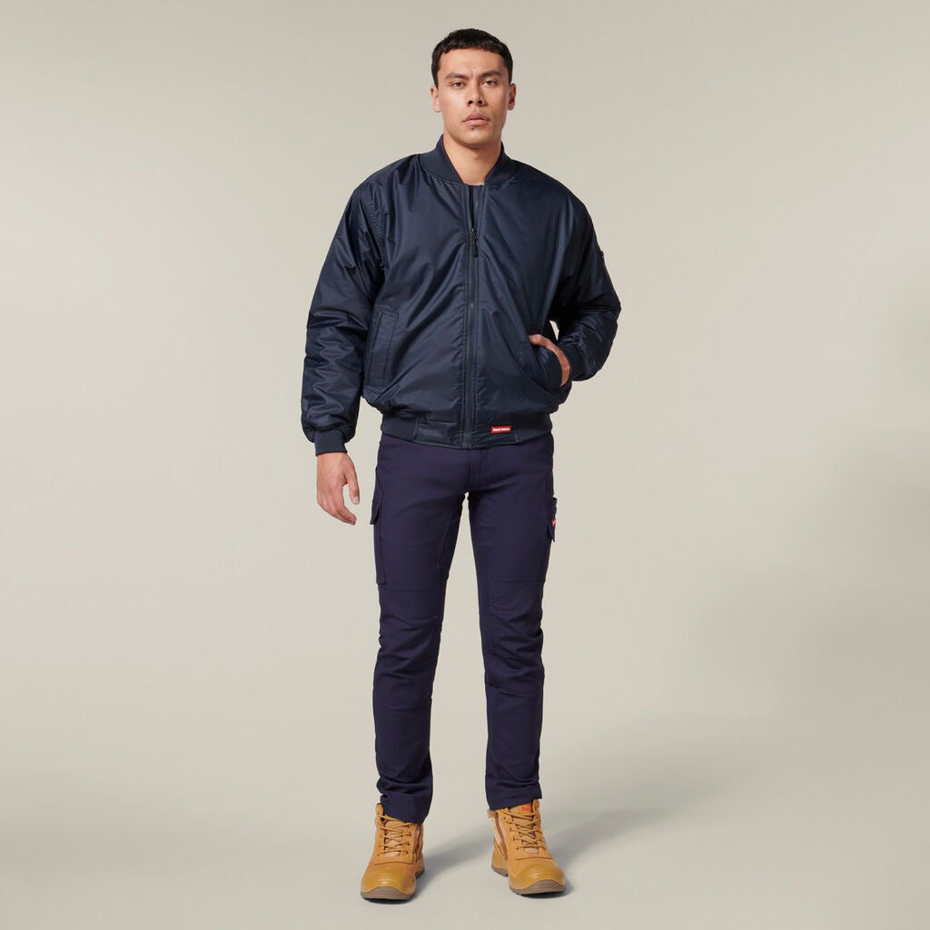 A person dressed in a navy bomber jacket, dark pants, and tan work boots casually holds an Emz A5 Notebook in one hand while keeping the other in their jacket pocket against a light beige backdrop.