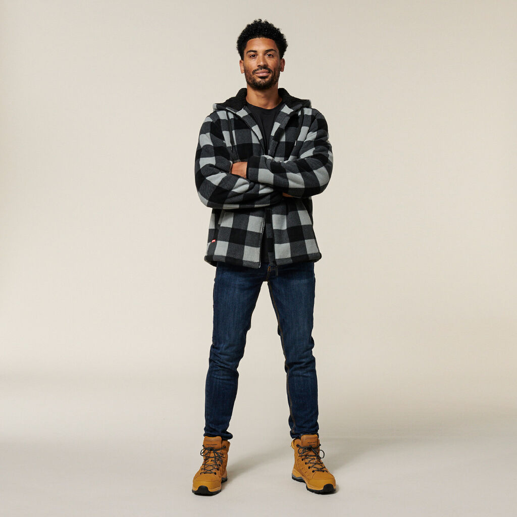 A man stands confidently with arms crossed, donning a black and gray checkered jacket reminiscent of an Emz A5 Notebook sketch, complemented by dark jeans and tan hiking boots, set against a neutral background.