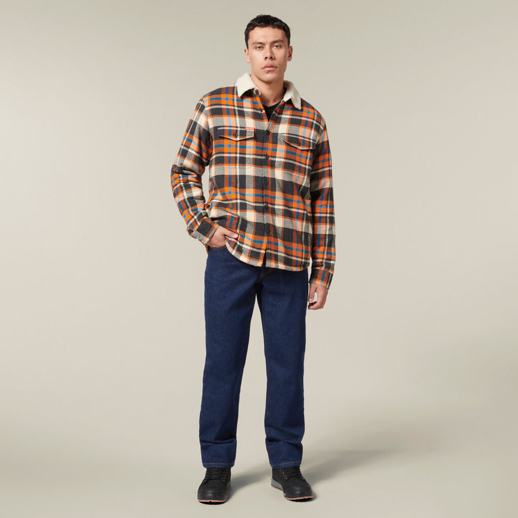 A man stands against a plain background holding an Emz A5 Notebook. Hes wearing a plaid flannel shirt with a shearling collar, blue pants, and black shoes. His short hair complements his casual pose with one hand in his pocket.