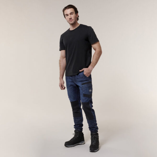 A man poses confidently against a simple backdrop, wearing a black t-shirt, dark blue cargo pants with black knee patches, and black boots. He holds an Emz A5 Notebook in his left hand while his right hand rests in his pocket.