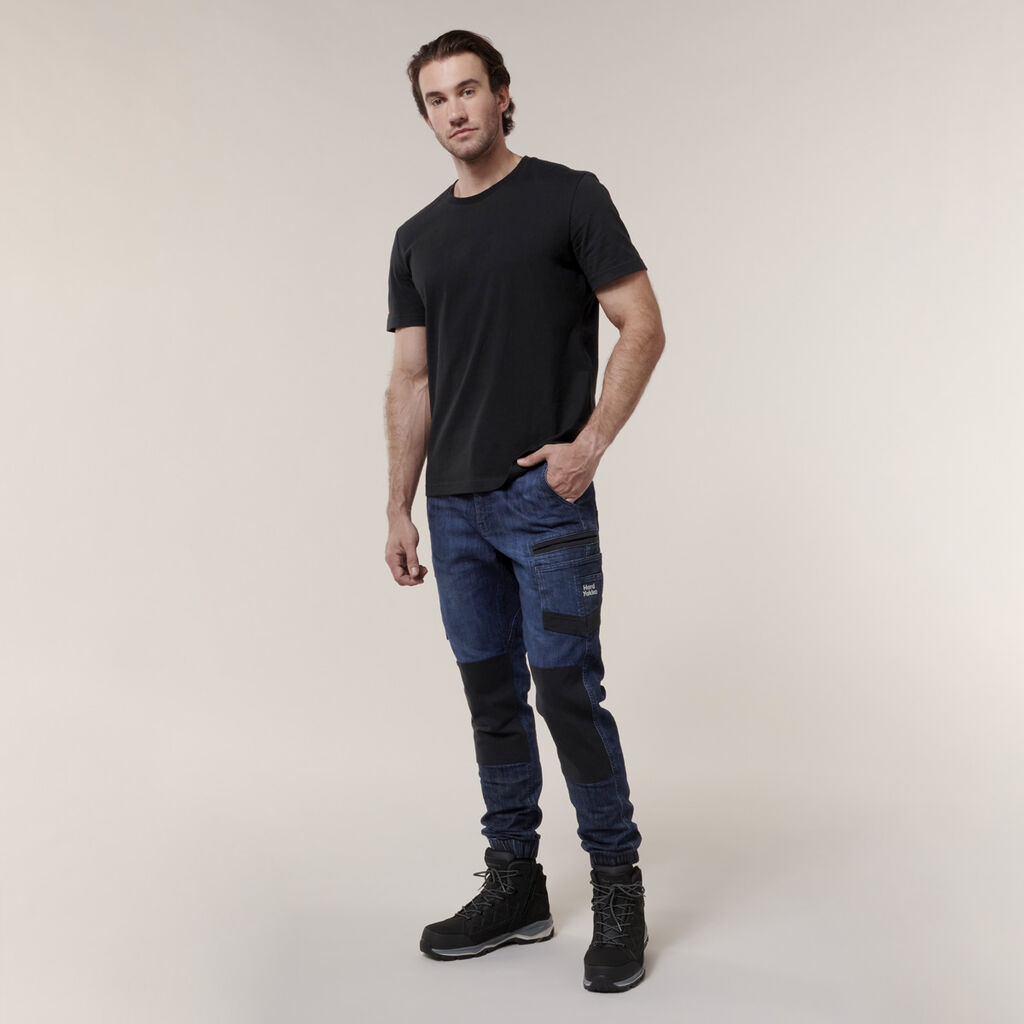 A man poses confidently against a simple backdrop, wearing a black t-shirt, dark blue cargo pants with black knee patches, and black boots. He holds an Emz A5 Notebook in his left hand while his right hand rests in his pocket.